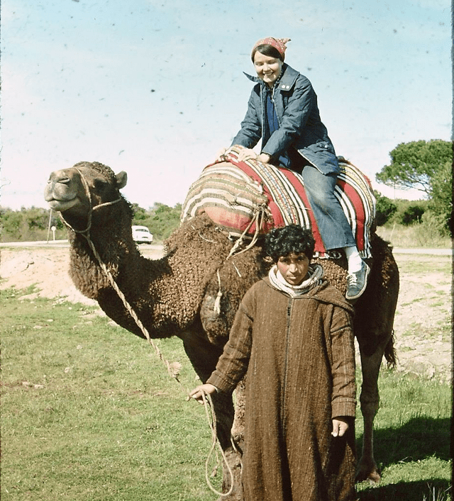 older photo of woman sitting on camel with young man standing beside her hold the rein